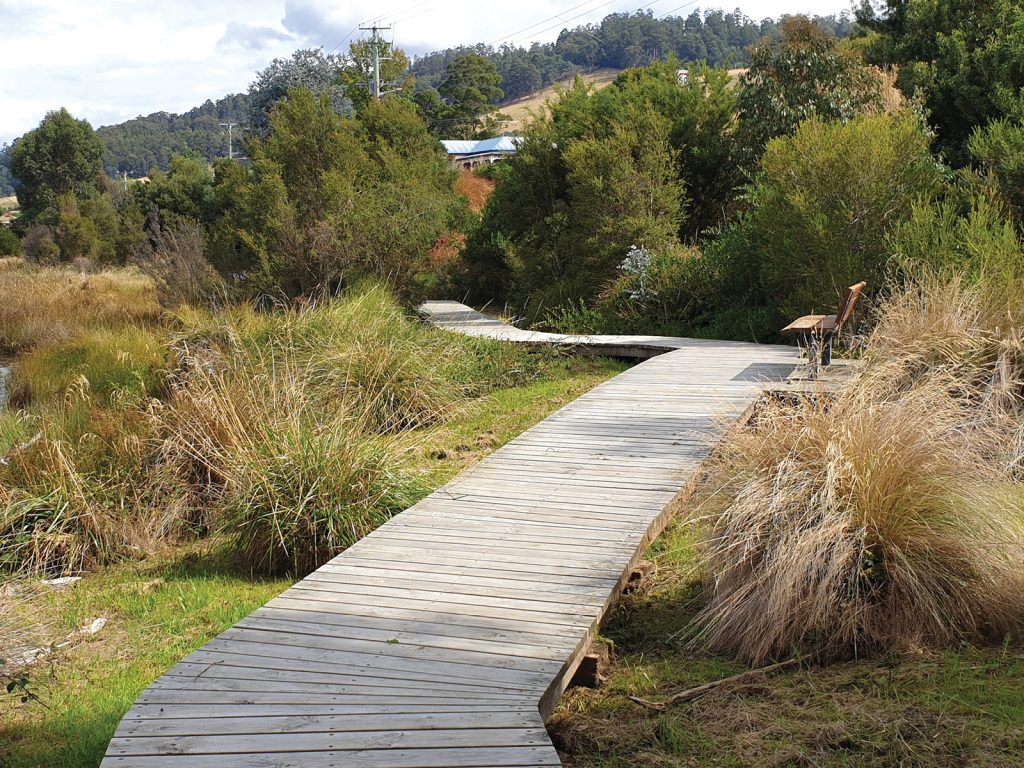 Wooden boardwalk curving through dense greenery with tall grass on both sides, leading towards a bench. Hills and a building are visible in the background under a partly cloudy sky.