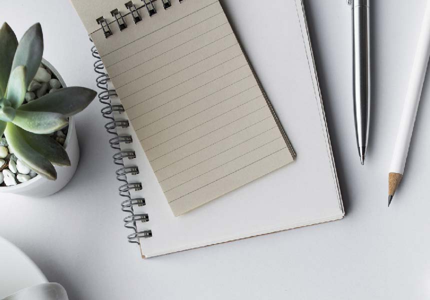 A top view of a desk with a spiral notebook and a lined notepad on top. Theres a silver pen, a white pencil, and a small potted succulent nearby on a white surface.