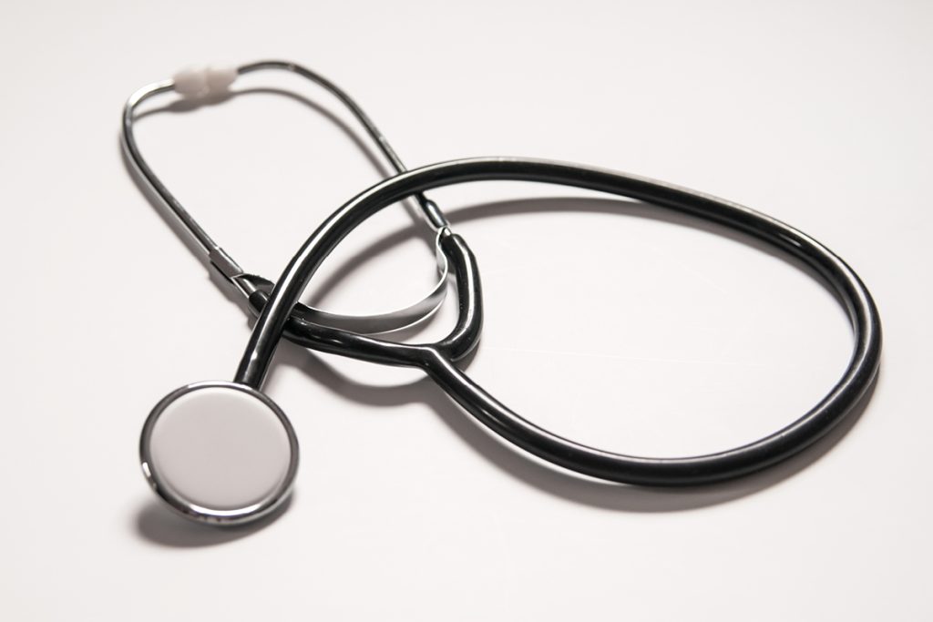 A stethoscope with a black tube and metallic ends is placed on a white surface. The chest piece is visible in the foreground, while the ear tubes and earpieces are slightly out of focus in the background.