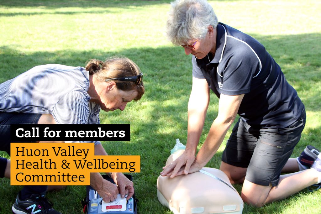 Two people perform CPR on a mannequin outdoors on grass. One is compressing the chest while the other prepares an AED. Text overlay reads, Call for members Huon Valley Health & Wellbeing Committee.