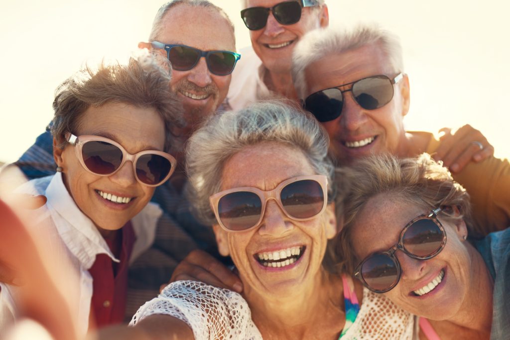 A group of six older adults wearing sunglasses smile joyfully at the camera while taking a selfie outdoors on a sunny day. They appear to be enjoying themselves, with clear blue skies in the background.