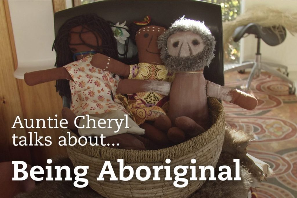 Three handmade dolls dressed in colorful patterns sit in a woven basket on a fur surface. The text overlay reads, Auntie Cheryl talks about... Being Aboriginal. The background features a sunny room with a chair and rug.