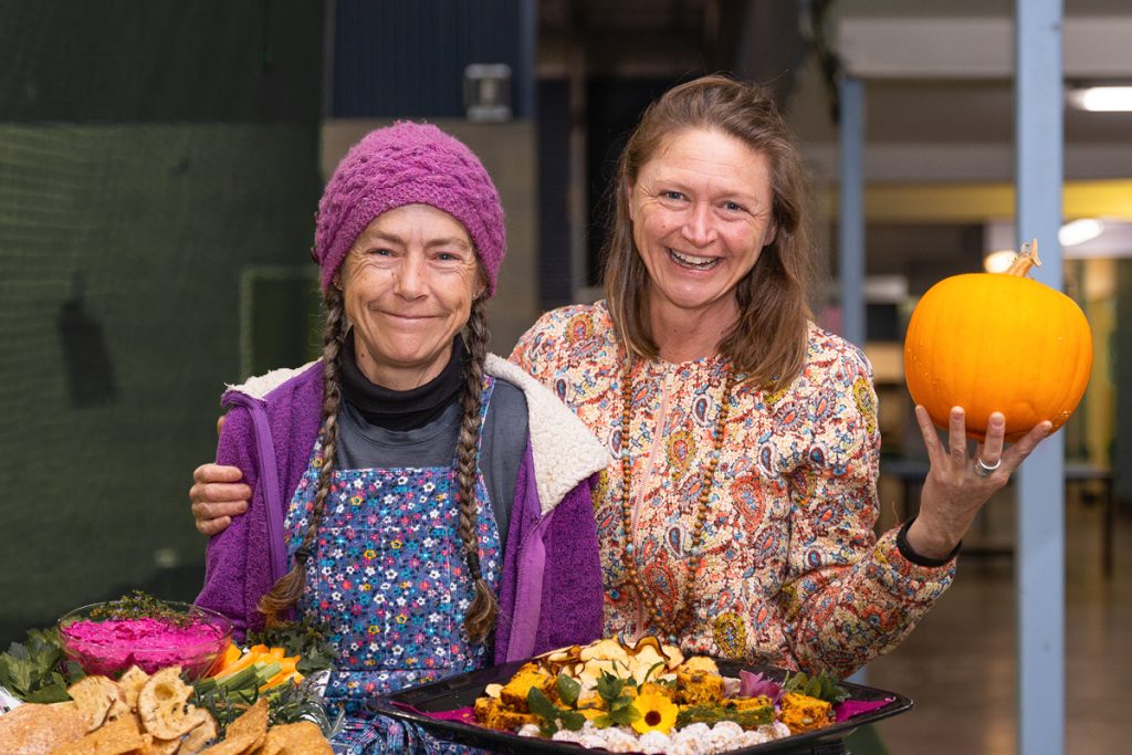 Two women smiling indoors, one with a purple beanie and braids holding a tray of food with colorful spreads, the other with brown hair holding a pumpkin. They stand close together in a cheerful setting.