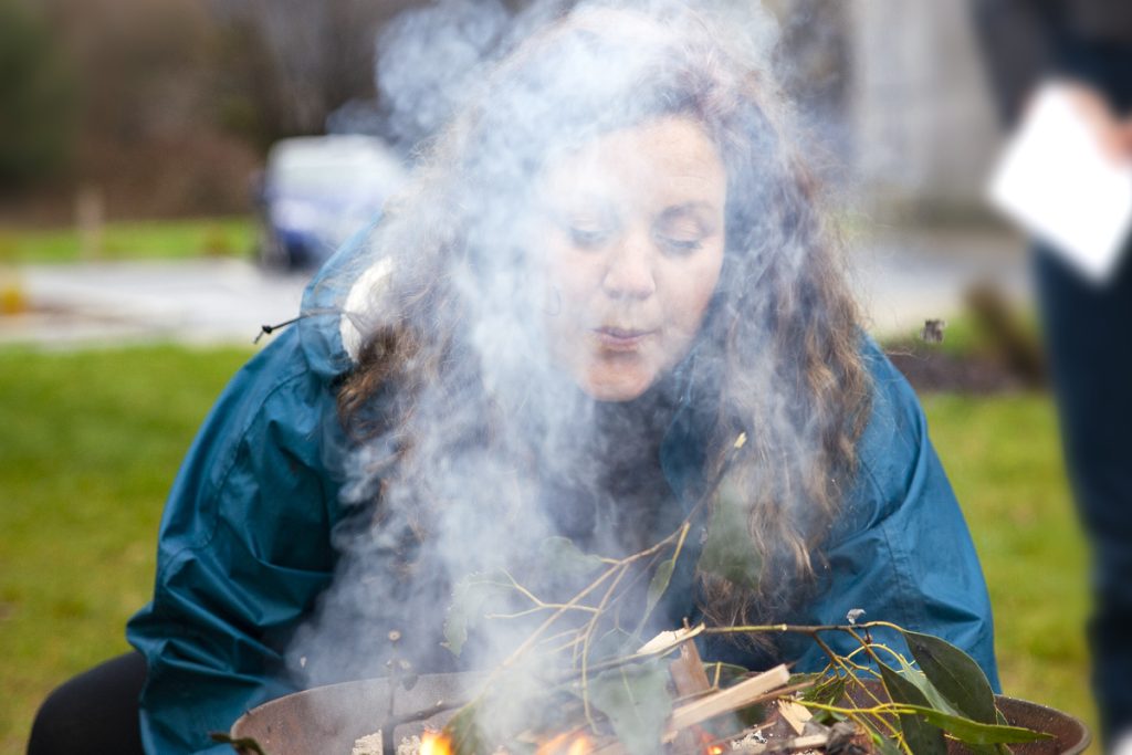 A woman with curly hair wearing a teal jacket blows into a smoky fire pit. The surrounding area is grassy, with blurred trees and a parked car in the background. The scene looks outdoors and somewhat overcast.