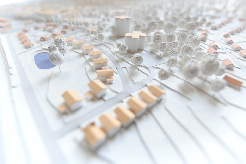 A miniature architectural model of a suburban neighborhood, featuring rows of small white and brown houses, a blue pond, trees, and three prominent white tower-like structures with cross-shaped tops. The scene is softly lit.