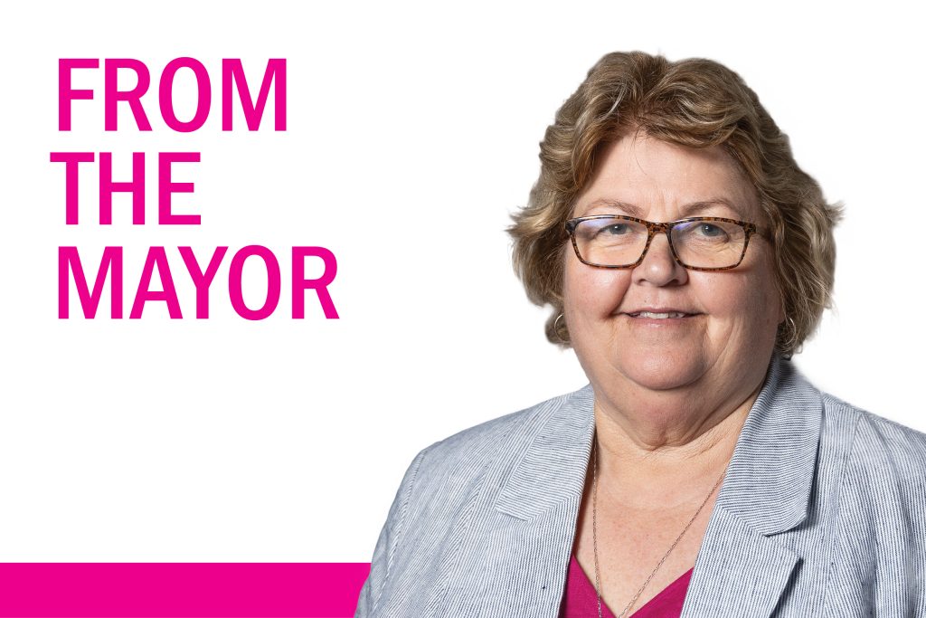 A person with glasses and short hair wearing a light gray blazer over a pink top stands smiling. The words FROM THE MAYOR are in bold pink letters on the left against a white background.