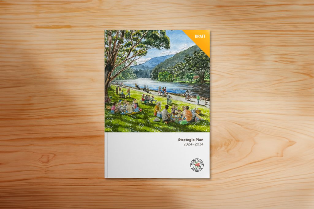 A draft strategic plan for 2024-2034 with a cover image depicting people relaxing by a river in a park, surrounded by trees and mountains under a clear sky. The document rests on a wooden surface.