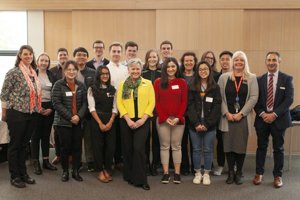 A group of people posing for a photo in a room with wooden walls. They are standing in two rows and smiling at the camera. Some are wearing name tags and ID lanyards. The group includes a mix of ages and genders.