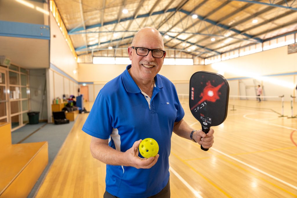 A man in a blue shirt holding a pickleball paddle and a yellow ball smiles at an indoor sports court. Behind him, people are playing on a wooden floor under a high ceiling with skylights.
