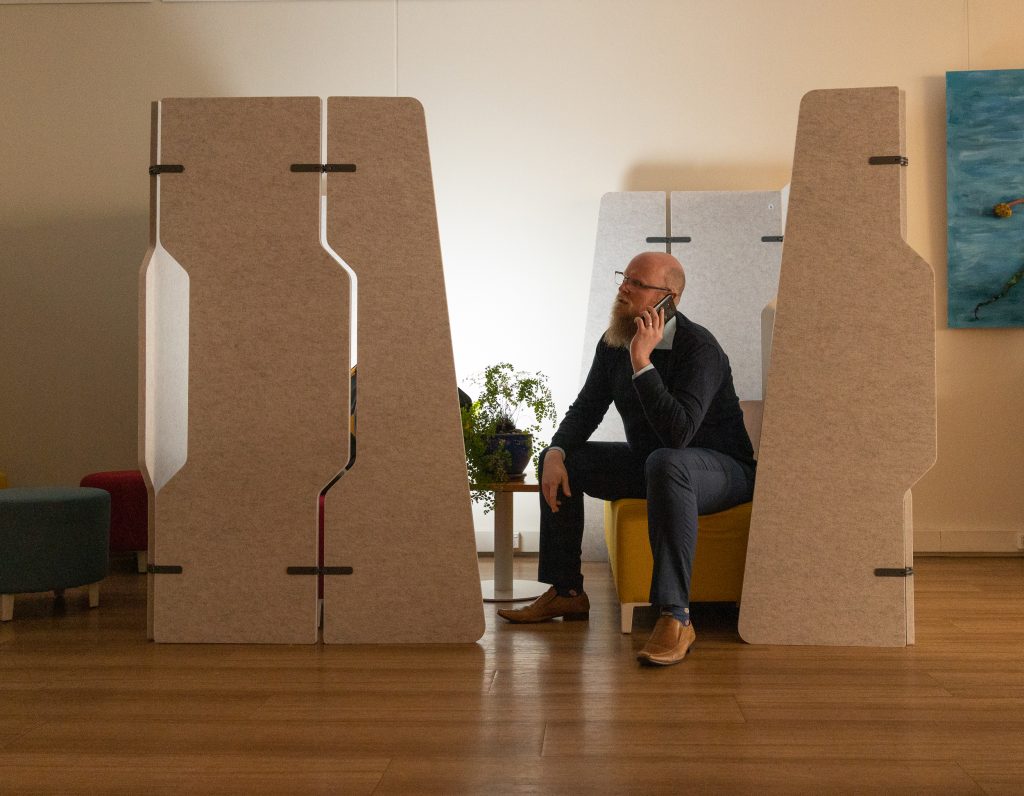 A person with a beard sits in a modern, gray office booth, talking on a smartphone. They are wearing a dark sweater and jeans. There are plants nearby and an abstract painting on the wall. The floor is wooden.