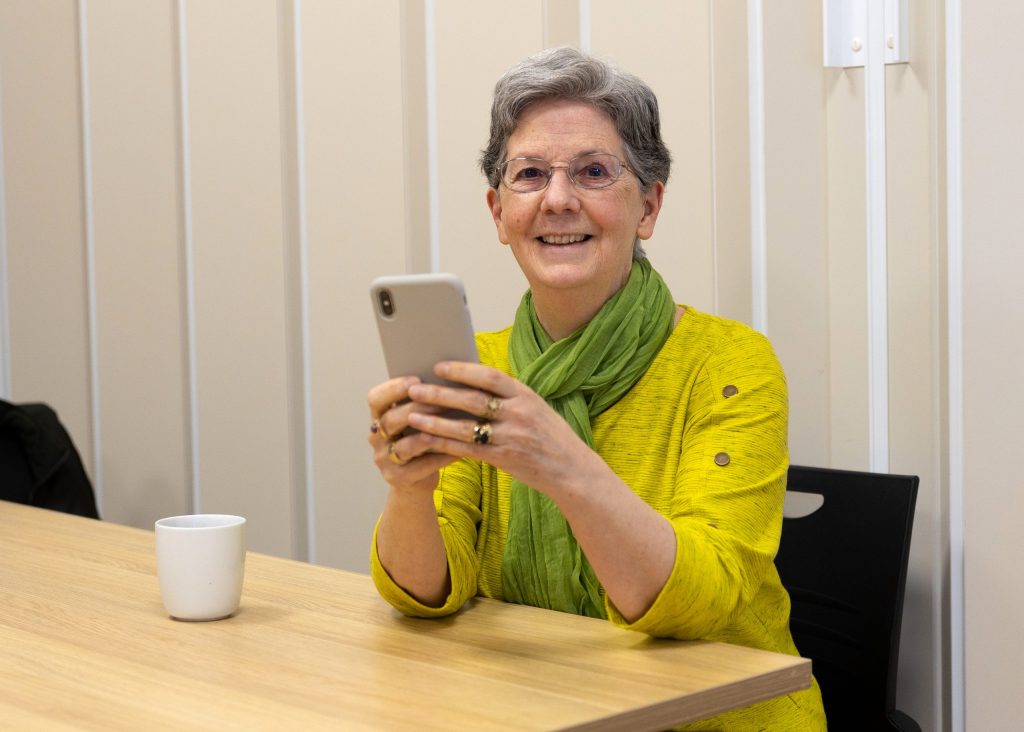 Elderly woman with short gray hair and glasses, wearing a yellow shirt and green scarf, sits at a wooden table holding a smartphone. A white mug is on the table in front of her. She is smiling and looking ahead.