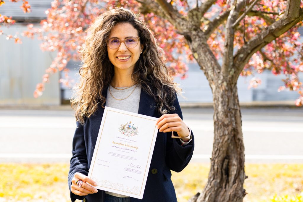 A woman with curly hair and glasses is smiling and holding a certificate outdoors. There is a tree with reddish leaves in the background. She is wearing a dark jacket and standing on a grassy area.