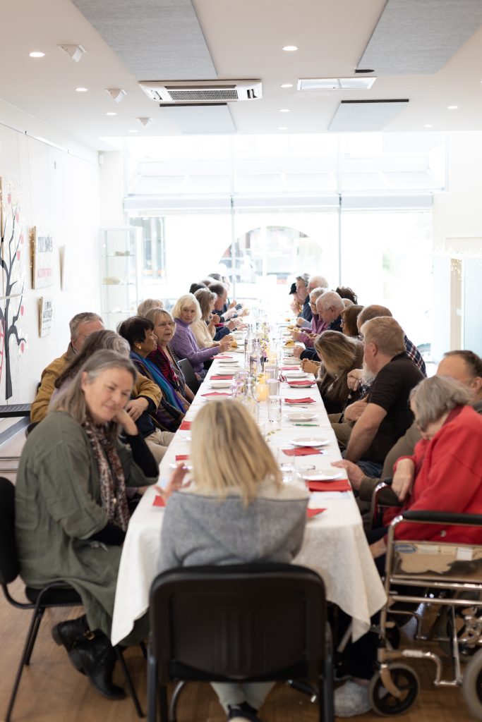 A diverse group of people sit together at a long, white-clothed table in a brightly lit room. They are engaged in conversation, with food and drinks on the table. Paintings are visible on the walls in the background.