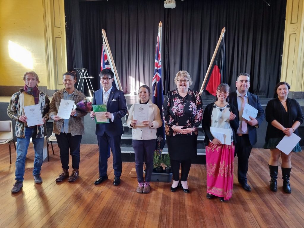 A group of seven people stand in a row, holding certificates, posing for a photo indoors. Behind them are two flags on display. They are standing on a wooden floor and the background features a black curtain.
