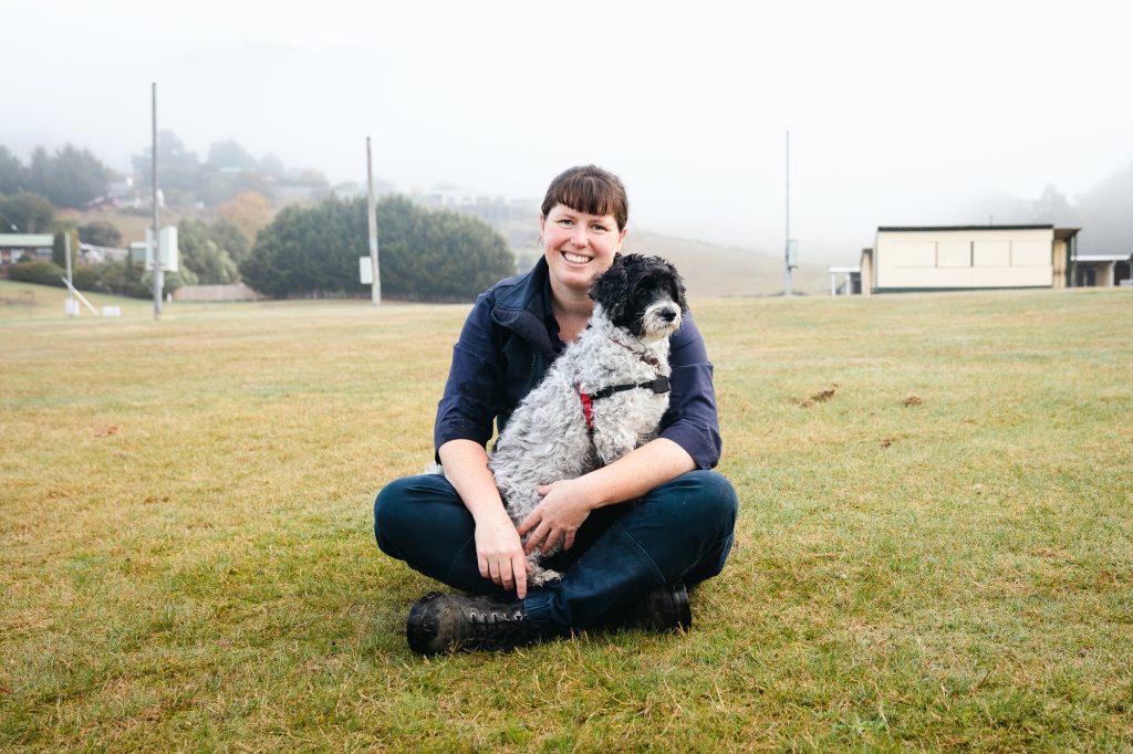 A person sitting cross-legged on grass, holding a small black and white dog. Theyre outdoors on a foggy day, with hills and trees in the background. The person is smiling and dressed in a blue top and boots.