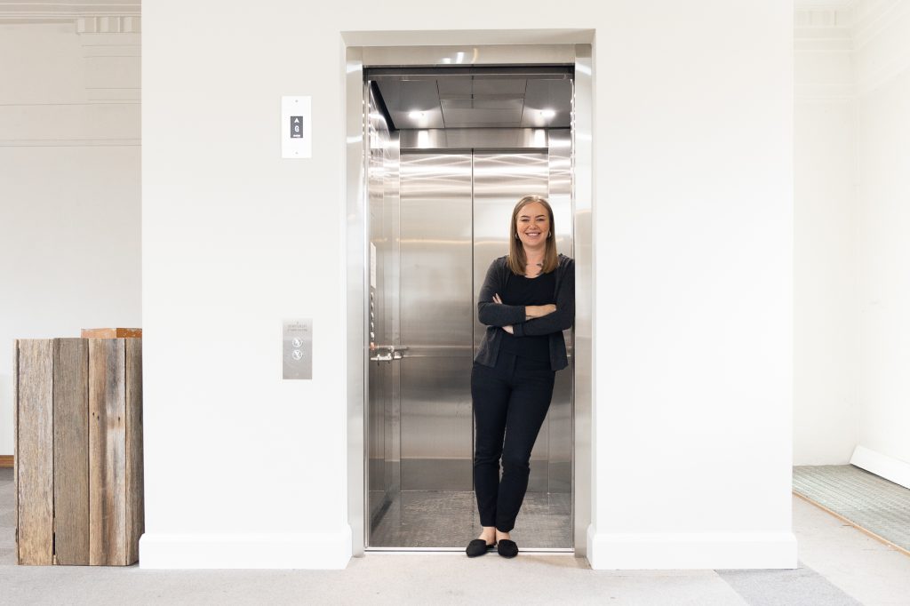 A person stands in the doorway of an open elevator, smiling and leaning against the door frame with arms crossed. The elevator is sleek and modern, and the surrounding walls are painted white. A stack of wooden boards is on the left.