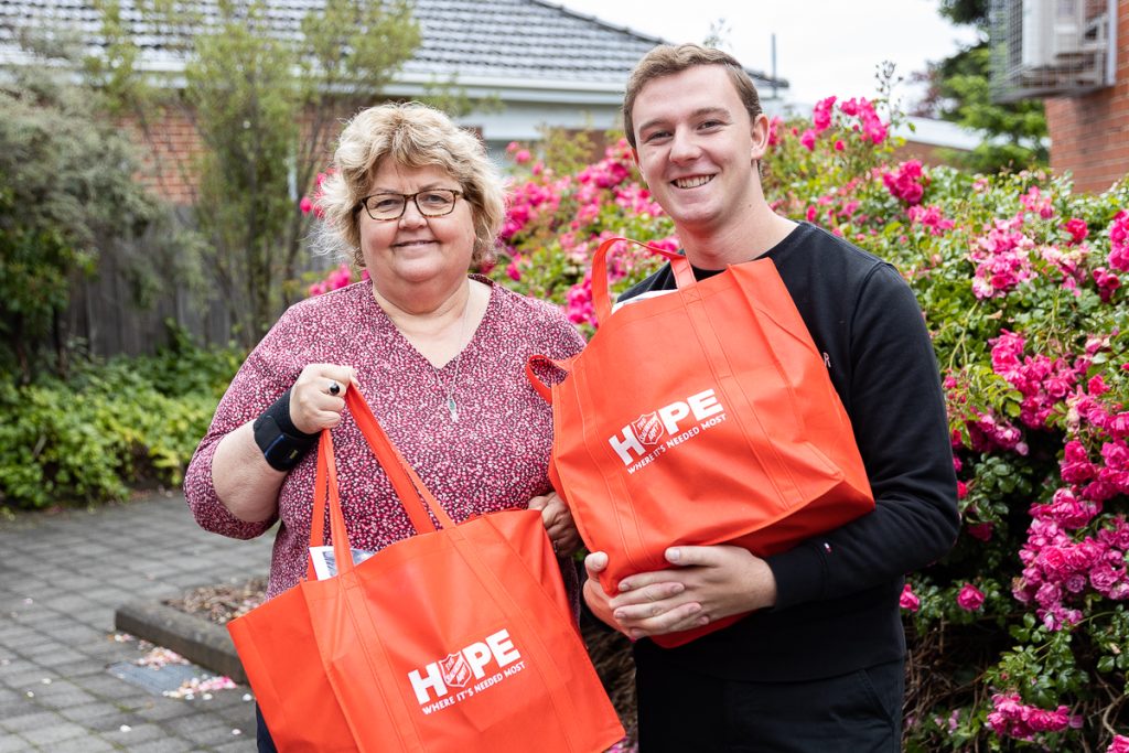 Two people are smiling and holding bright orange bags labeled Hope. They are standing on a paved path with vibrant pink flowers and a brick building in the background.