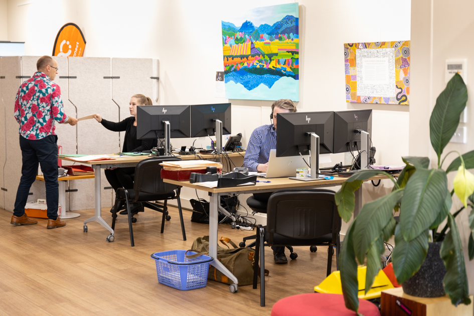 An office scene with three people working. A man in a floral shirt talks to a woman at a computer. Another person is focused on their laptop. The room has a large plant, colorful wall art, and three computer monitors on desks.