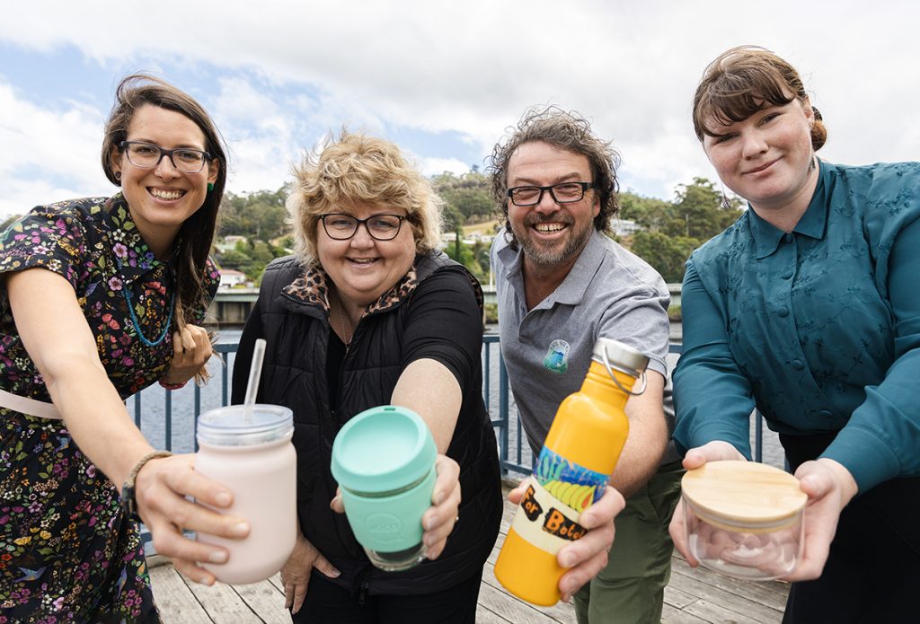 Four people standing on a wooden deck, smiling, and holding out reusable drink containers. The background shows trees and a partly cloudy sky. They appear to be promoting eco-friendly habits.