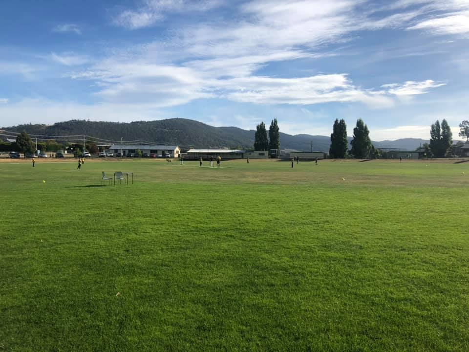 A wide grassy field under a blue sky with scattered clouds. In the distance, people are engaged in activities. Hills and trees line the horizon, and a few structures are visible near the fields edge.
