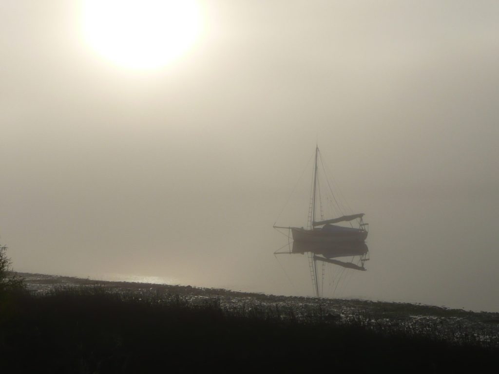 A sailboat is reflected on a calm, misty lake under a bright, hazy sun. The scene is serene and shrouded in fog, with the shoreline visible in the foreground.