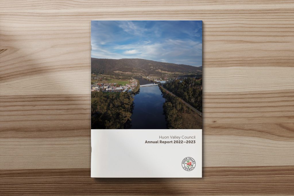 Cover of the Huon Valley Council Annual Report 2022-2023 on a wooden surface. Features an aerial view of a river flowing through a valley with hills and a town, under a partly cloudy sky. A small council logo is at the bottom.