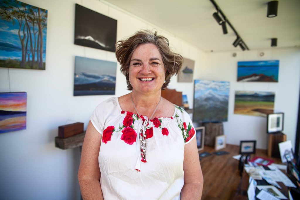 A smiling woman stands in an art gallery, wearing a white blouse with red floral embroidery. Paintings of landscapes adorn the walls around her, and various artworks are displayed on shelves and tables. The room has wooden flooring and bright lighting.