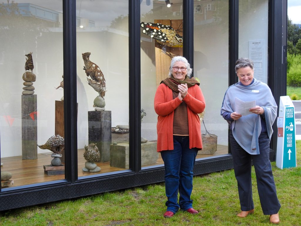 Two people stand outside a glass display with sculptures. One is holding a small sculpture, wearing a red sweater, glasses, and has gray hair. The other has short hair, a gray shirt, and is holding a paper. An exhibit of animal sculptures is visible behind them.