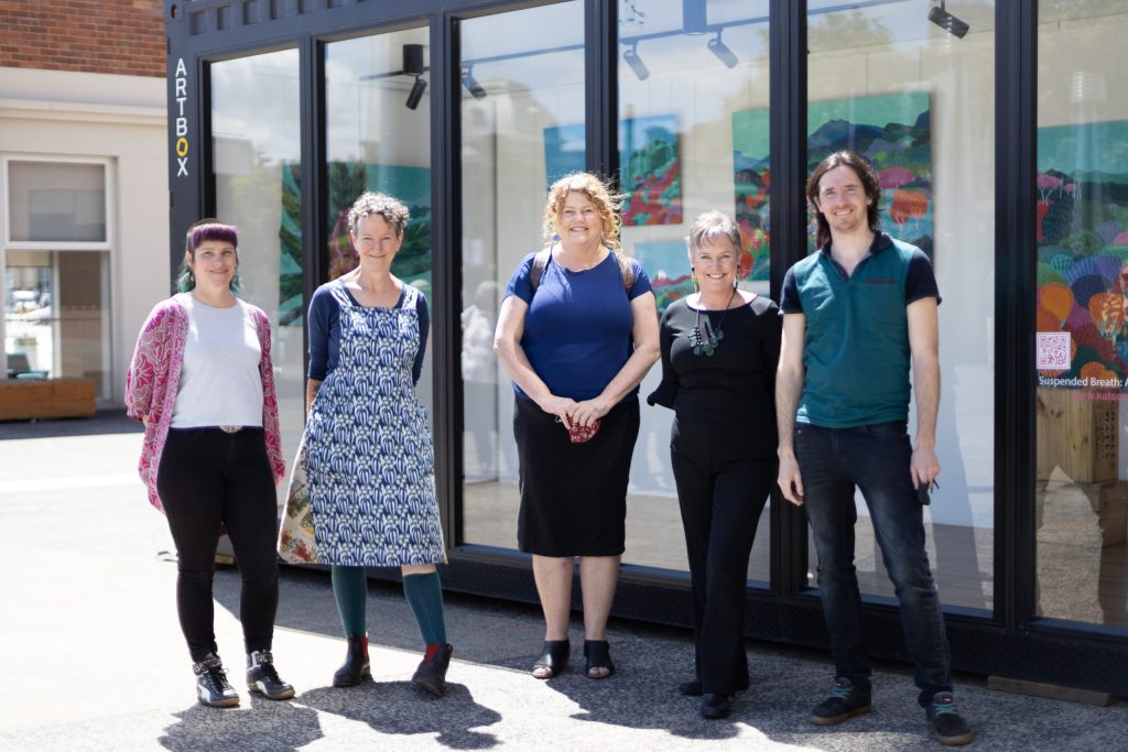 Five people stand smiling in front of a large art display window. A variety of colorful paintings are visible behind them. They are outdoors on a sunny day, and the group appears friendly and casual.