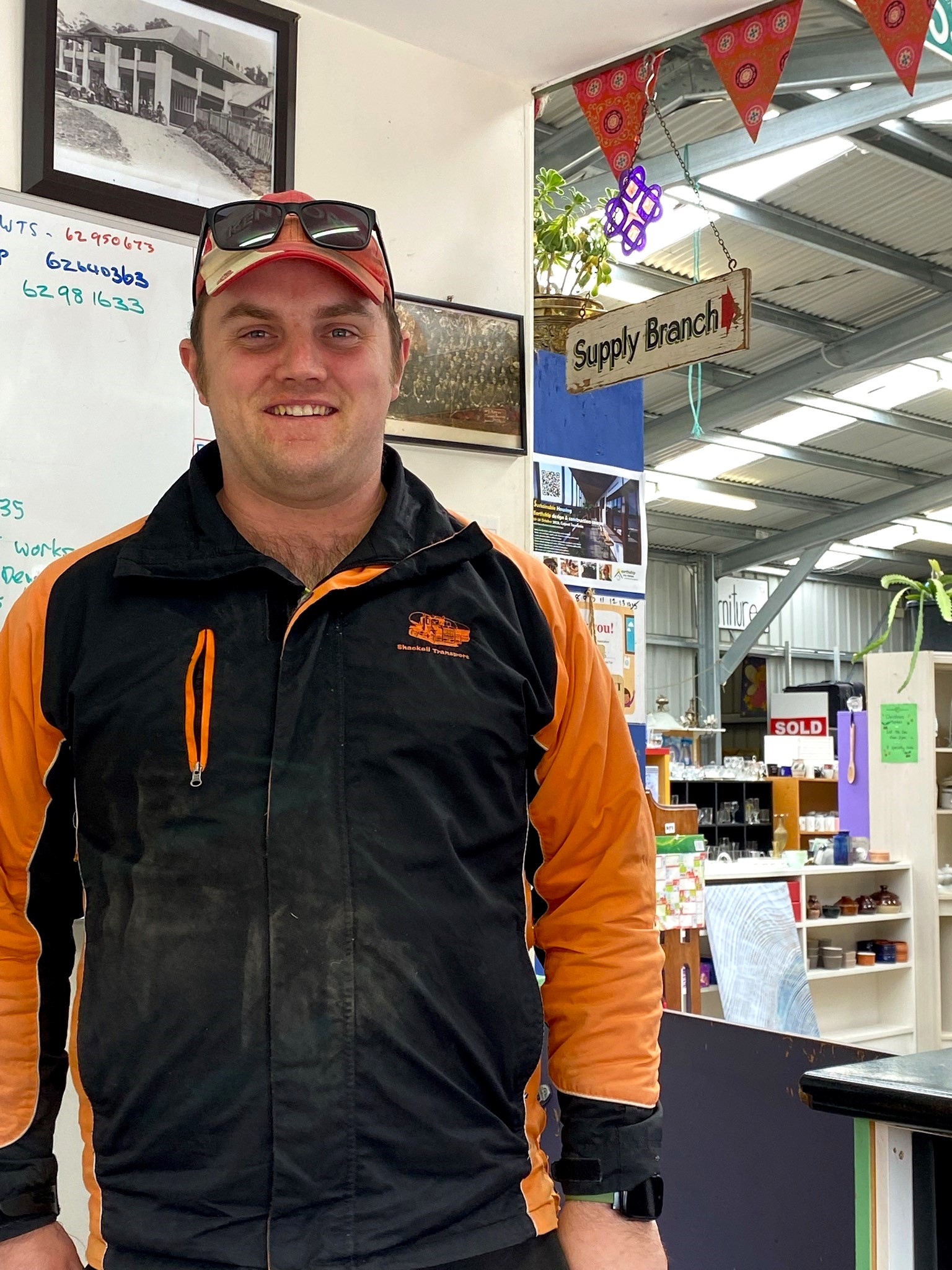 Man wearing a cap, sunglasses, and an orange and black jacket stands indoors. Behind him, a Supply Branch sign is visible, along with shelves and a whiteboard. The setting appears to be a store or warehouse with industrial decor.