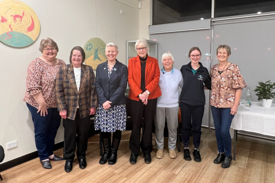 A group of seven women stand in a line indoors, smiling at the camera. They are dressed in a variety of outfits, from casual to formal. Behind them is a wall with artwork and a window with blinds. A small table with a plant is on the right.