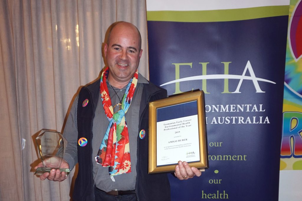 A person smiling and holding a glass trophy in their left hand and a framed certificate in their right hand. They are wearing a colorful scarf and buttons with rainbows. Behind them is a banner with Environmental Health Australia written on it.