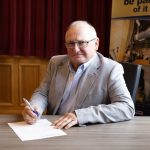 An older adult in a gray suit and glasses sits at a table, holding a pen over a document. Behind him are maroon curtains and a banner. A flag stands in the corner to the left.