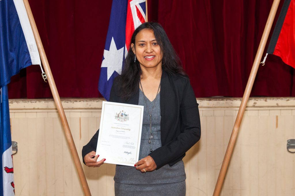 A woman in a gray dress and black blazer holds a certificate, standing in front of Australian flags and a red curtain.