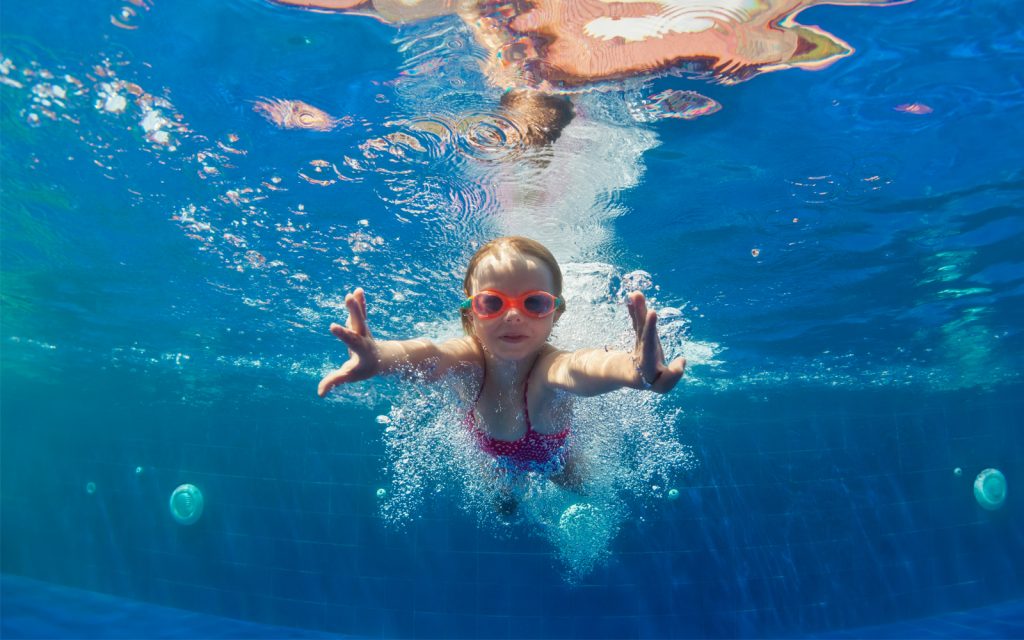 A young child wearing red goggles swims underwater in a pool, reaching forward with both hands. The blue water surrounds her, with light rippling on the surface above. Bubbles trail behind her as she moves confidently.