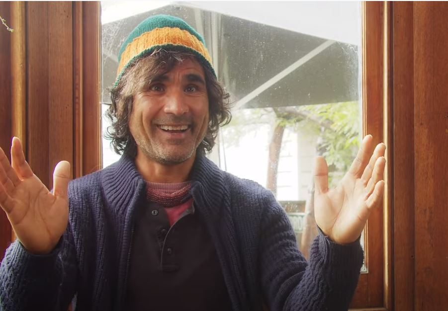 A person with long, wavy hair is wearing a colorful beanie and a blue sweater. They are smiling with their hands raised, seated in front of a window. The background shows some greenery outside.