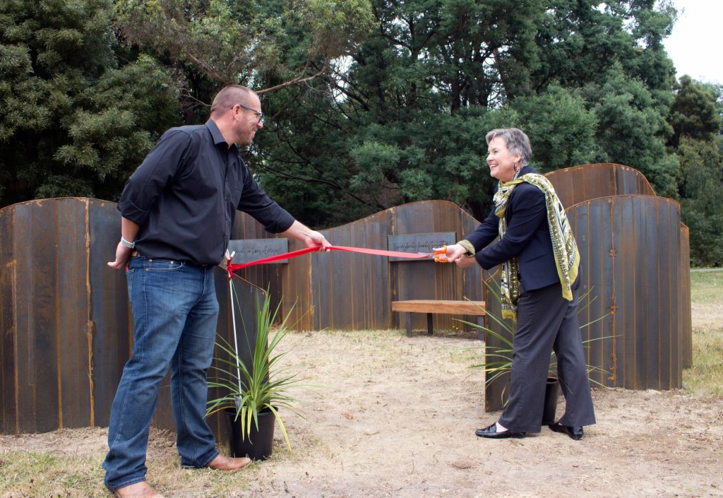 Two people are holding a red ribbon, preparing for a ribbon-cutting ceremony in an outdoor setting. Behind them is a curved, rust-colored metal structure, and in the foreground are two potted plants. Trees are visible in the background.