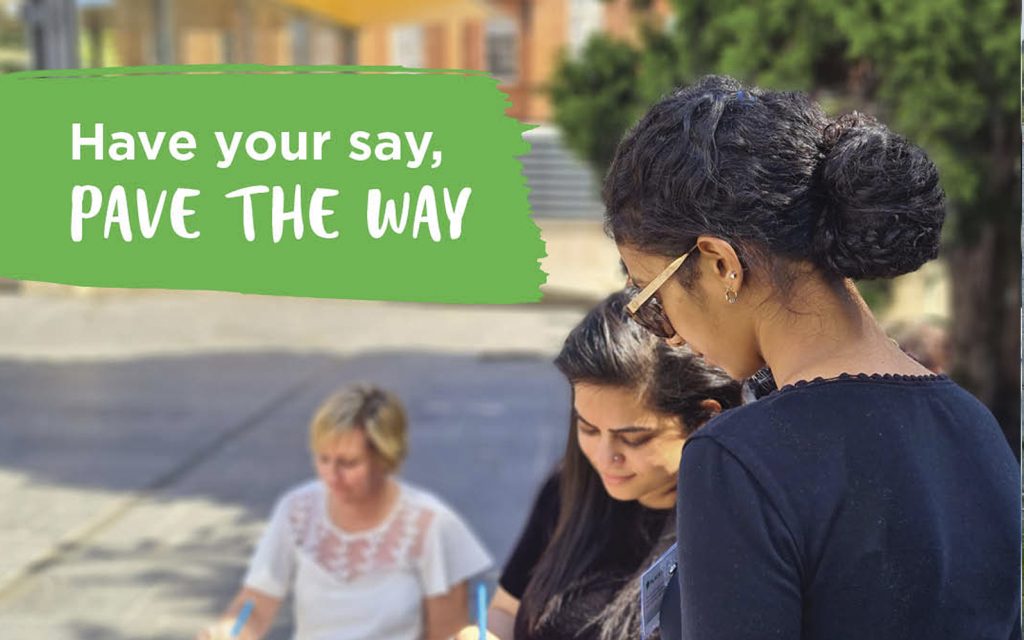 Three women are engaged in an outdoor activity. Two are focused on writing, while the third looks on. A green sign above them reads Have your say, PAVE THE WAY. Trees and buildings are visible in the background.