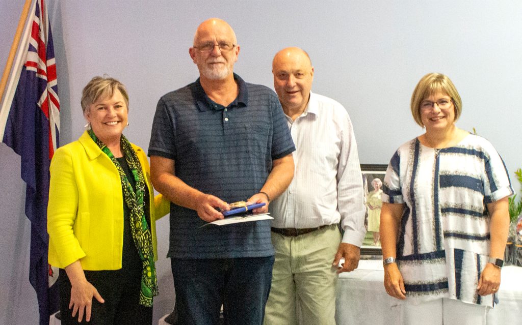 Four people standing side by side indoors, smiling. The man in the middle holds a small box. Theres an Australian flag in the background and a framed photo on a table. The group appears to be celebrating or commemorating an event.