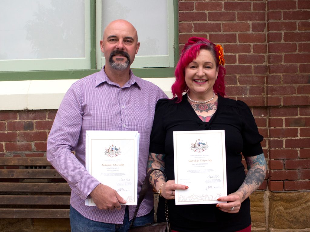 A man and a woman stand outside a brick building, each holding a certificate. The man wears a lavender shirt, and the woman has bright pink hair and tattoos. They are smiling, celebrating their Australian citizenship.