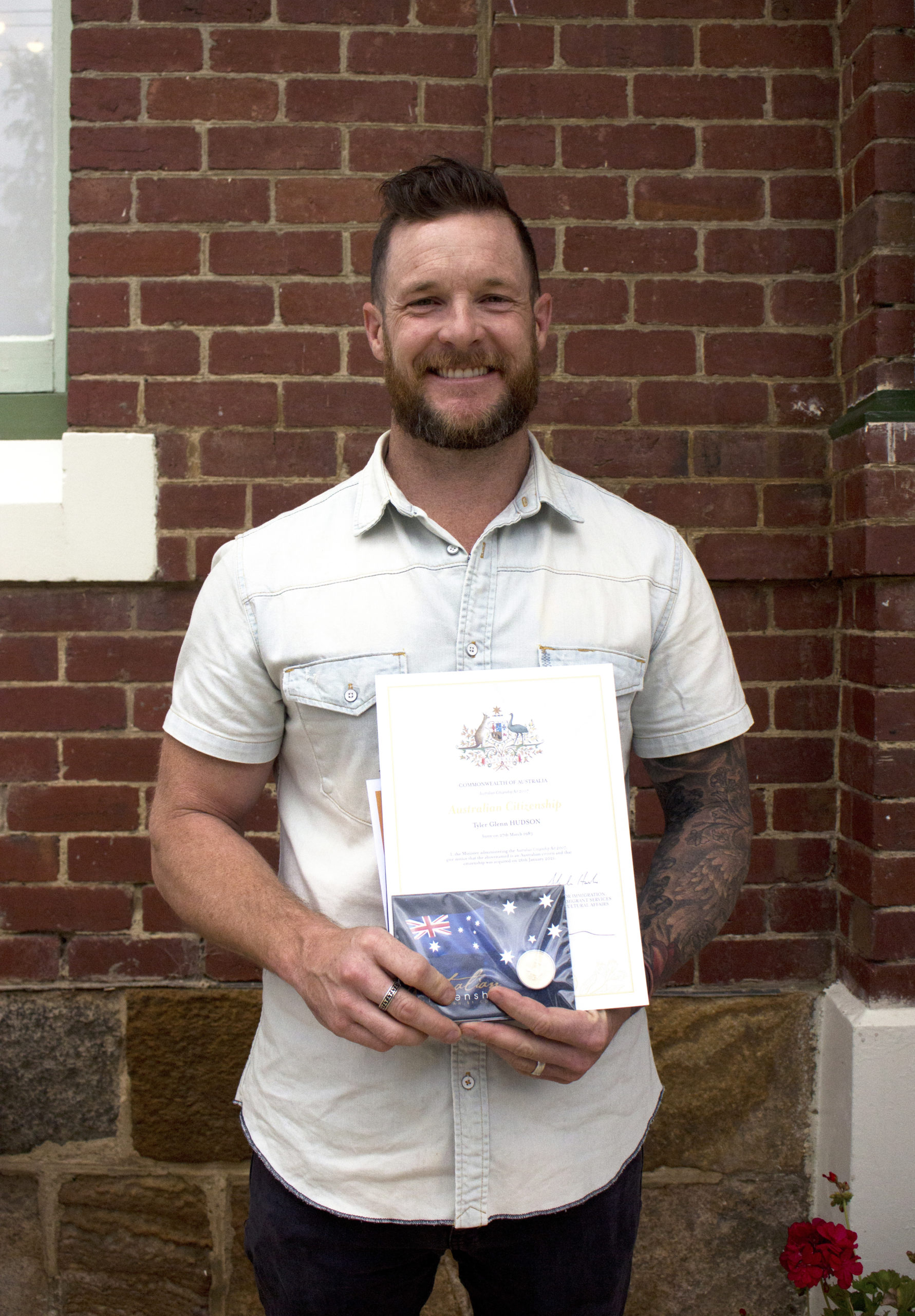 A man with a beard, wearing a light-colored shirt, holds a certificate and a small Australian flag standing in front of a brick wall. He is smiling.