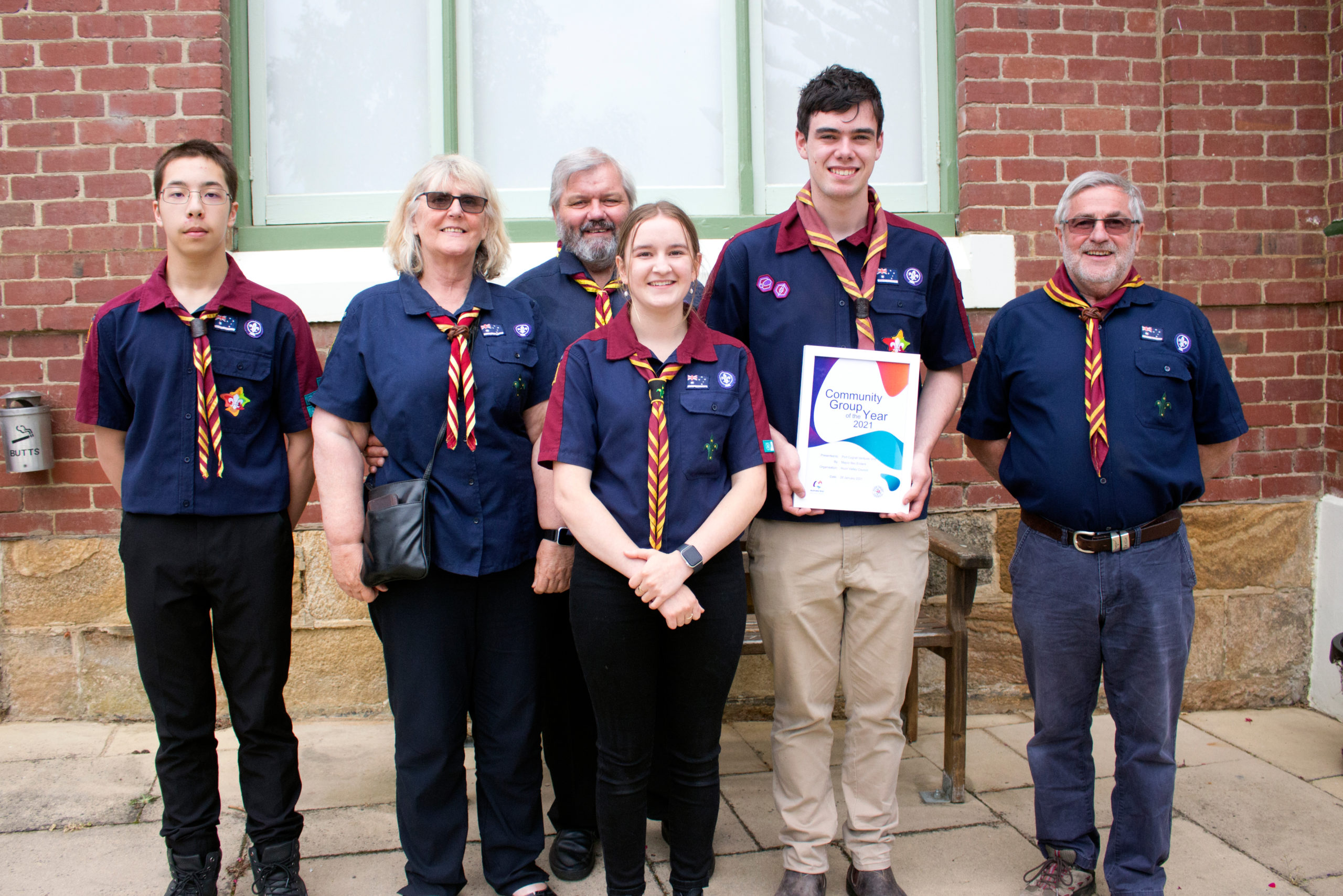 A group of six people in scout uniforms stands in front of a brick building. The person in the center holds a Community Contributor Award certificate. All are smiling, with trees and a large window in the background.