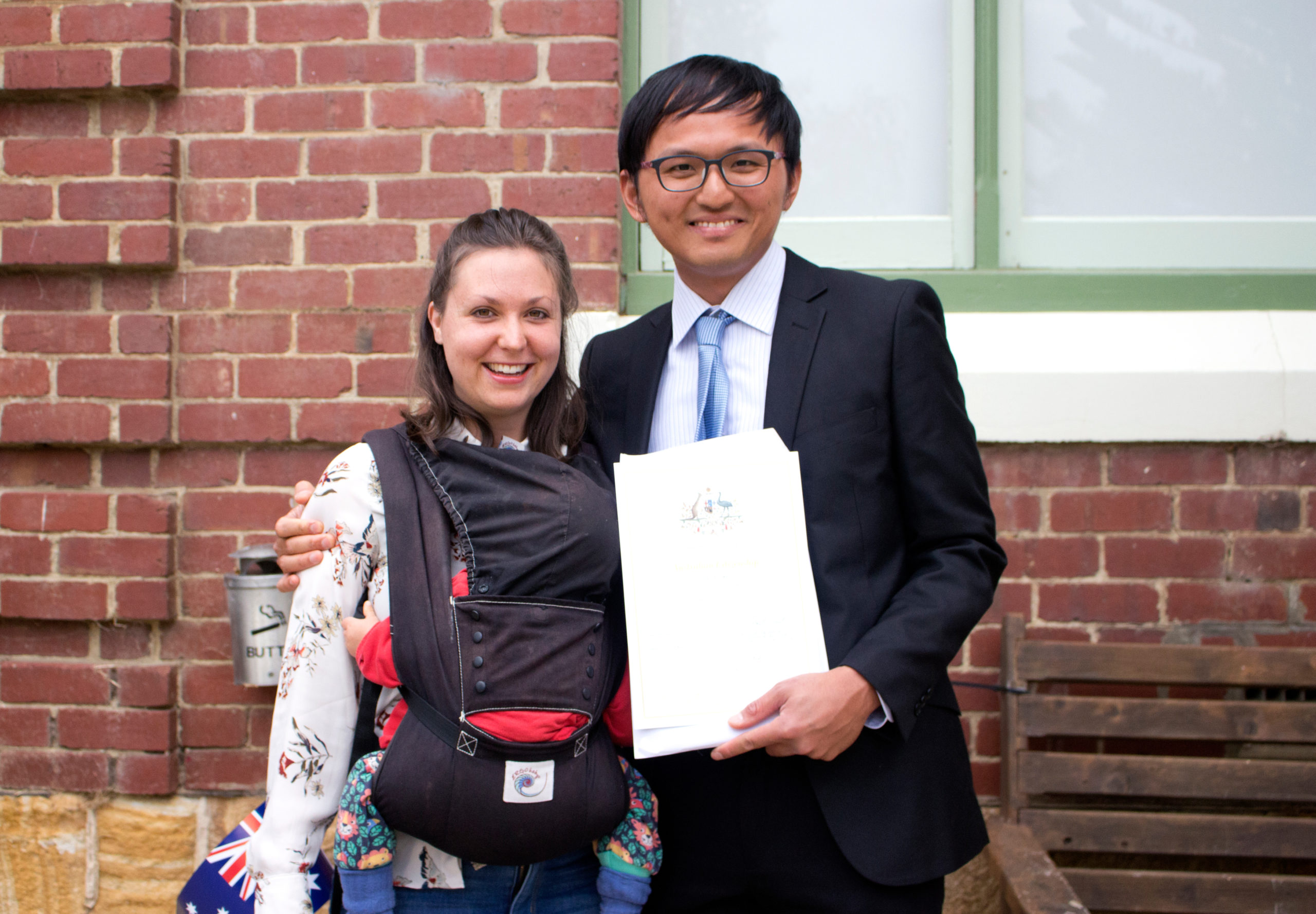 A smiling man in a suit holds a certificate and stands next to a woman holding a baby carrier. They pose in front of a brick building with a green-framed window. The womans outfit is patterned, and the mans tie is light blue.