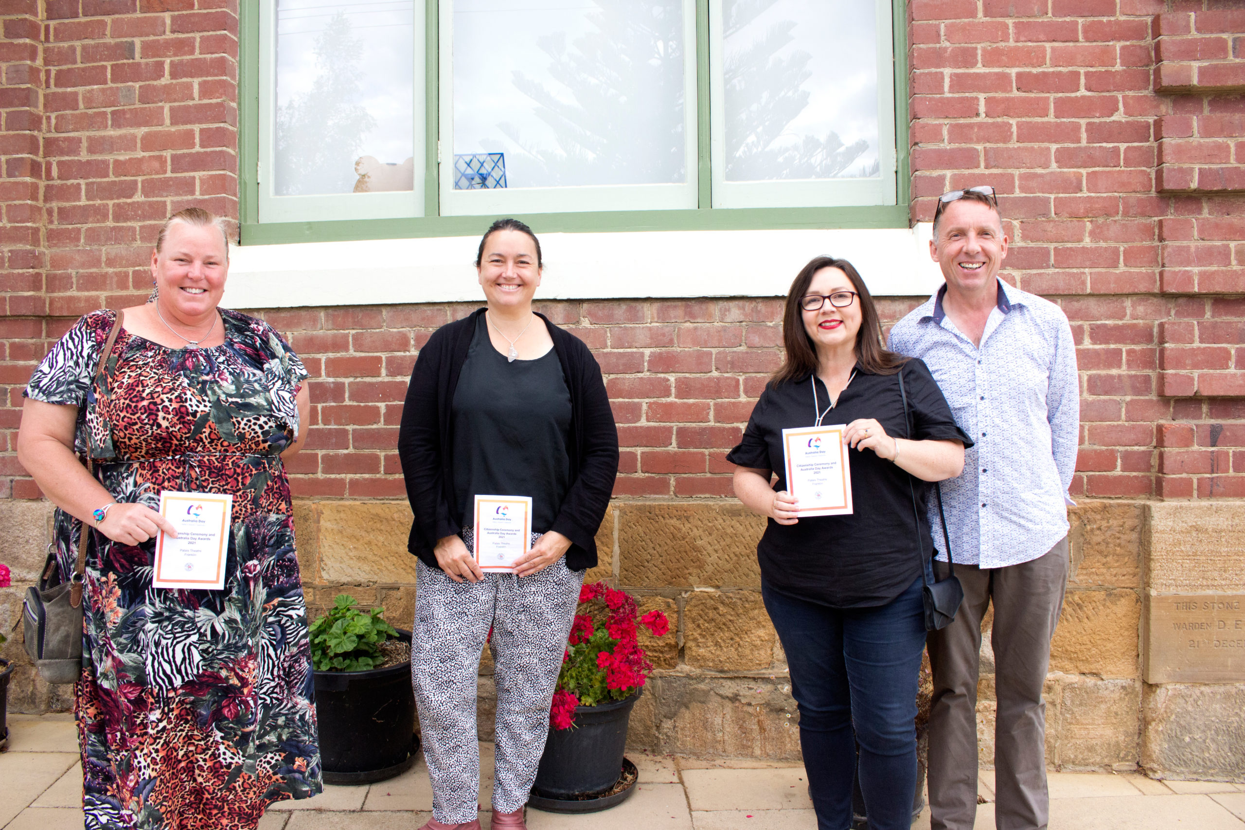 Four people smiling and holding certificates stand in front of a brick building. There are potted plants and a window behind them.