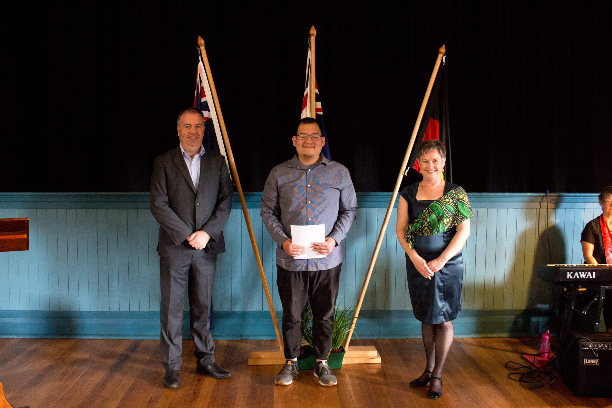 Three people stand smiling in front of crossed flags in a hall. The man in the center holds a certificate. A pianist is partially visible to the right. The backdrop includes blue paneling and wooden flooring.