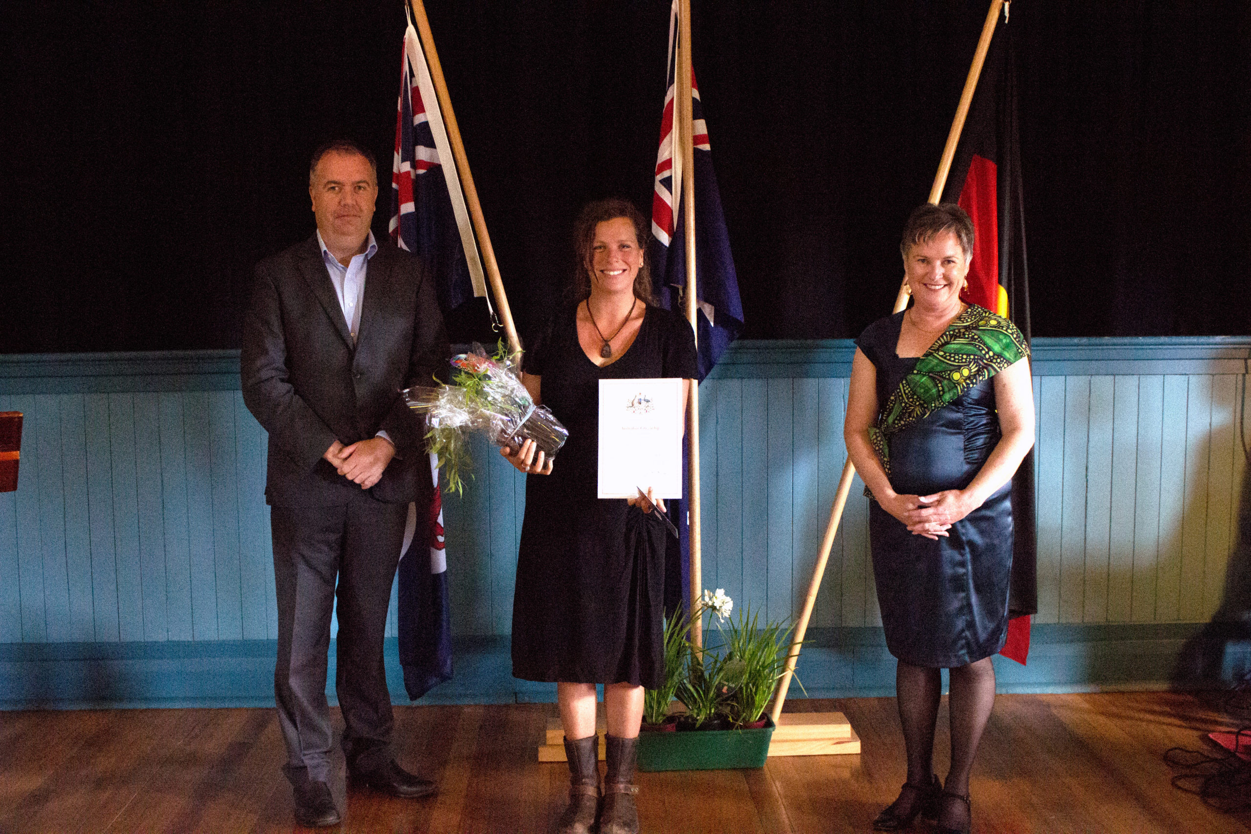 Three people are standing indoors. The person in the middle holds flowers and a certificate. They are flanked by two other people, one in a suit and the other in a dress. Three flags are displayed behind them.