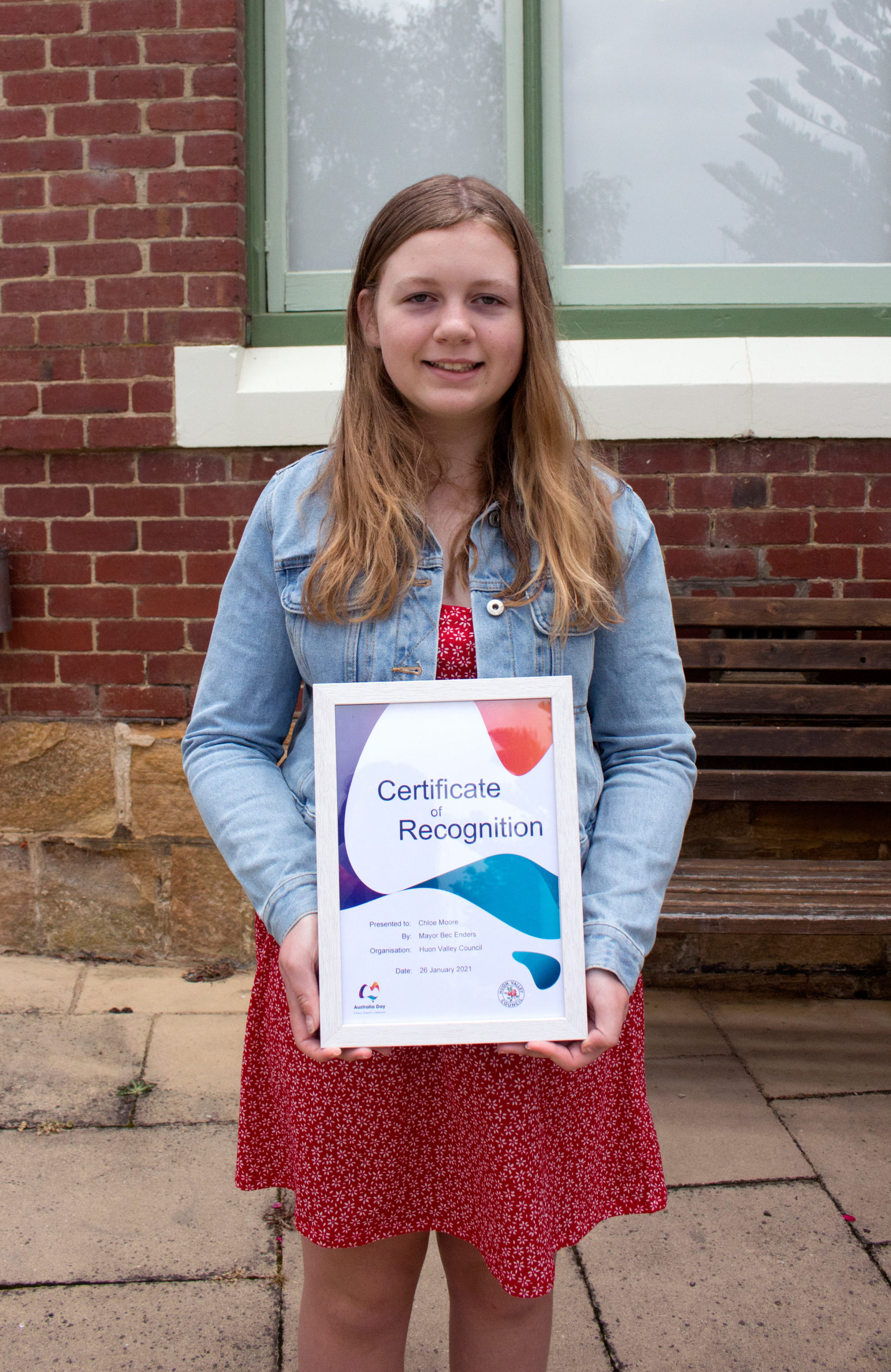 A young woman stands outside holding a Certificate of Recognition. She wears a red patterned dress and a denim jacket. The background includes a bench, a brick wall, and a window.