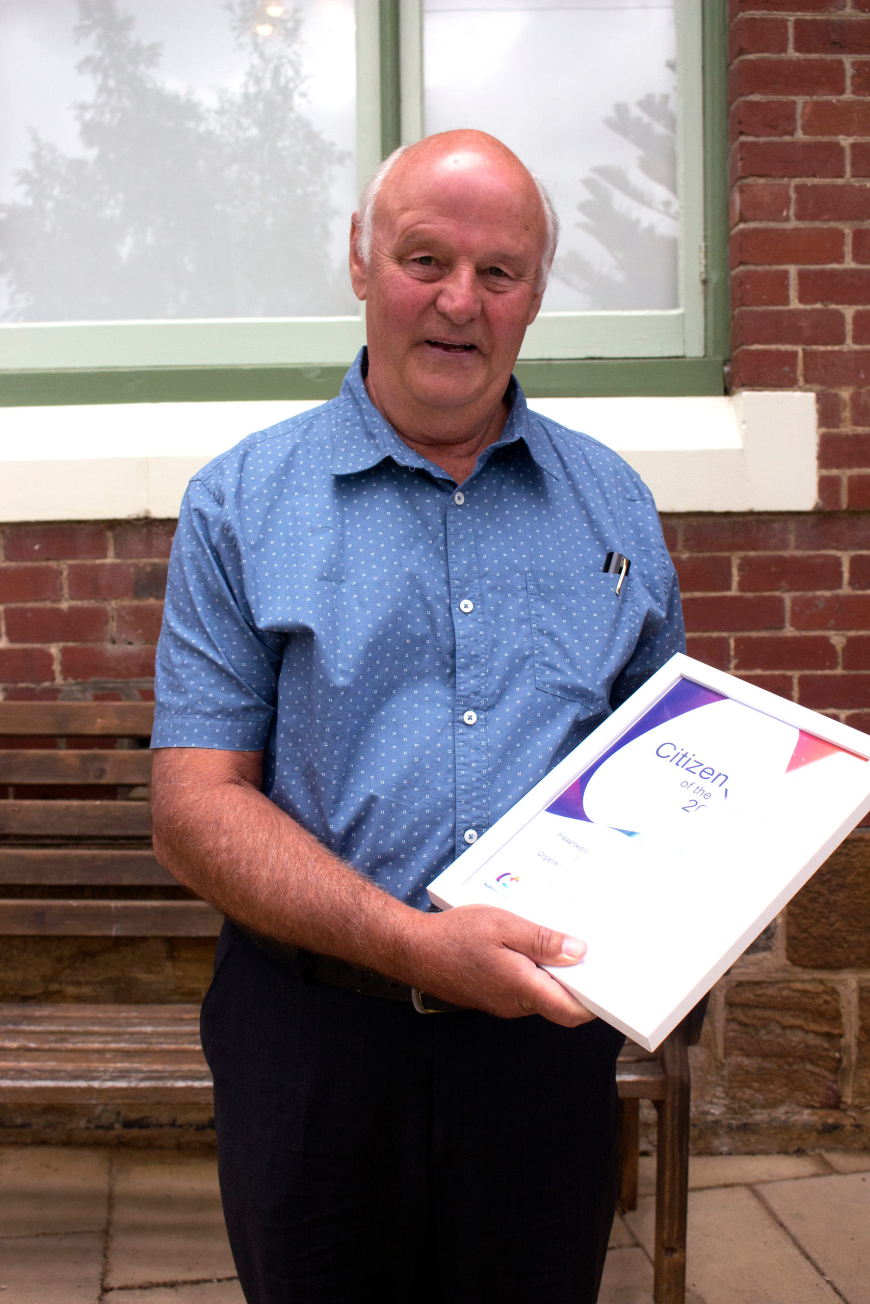 A man in a blue shirt stands in front of a window and brick wall, holding a framed certificate.