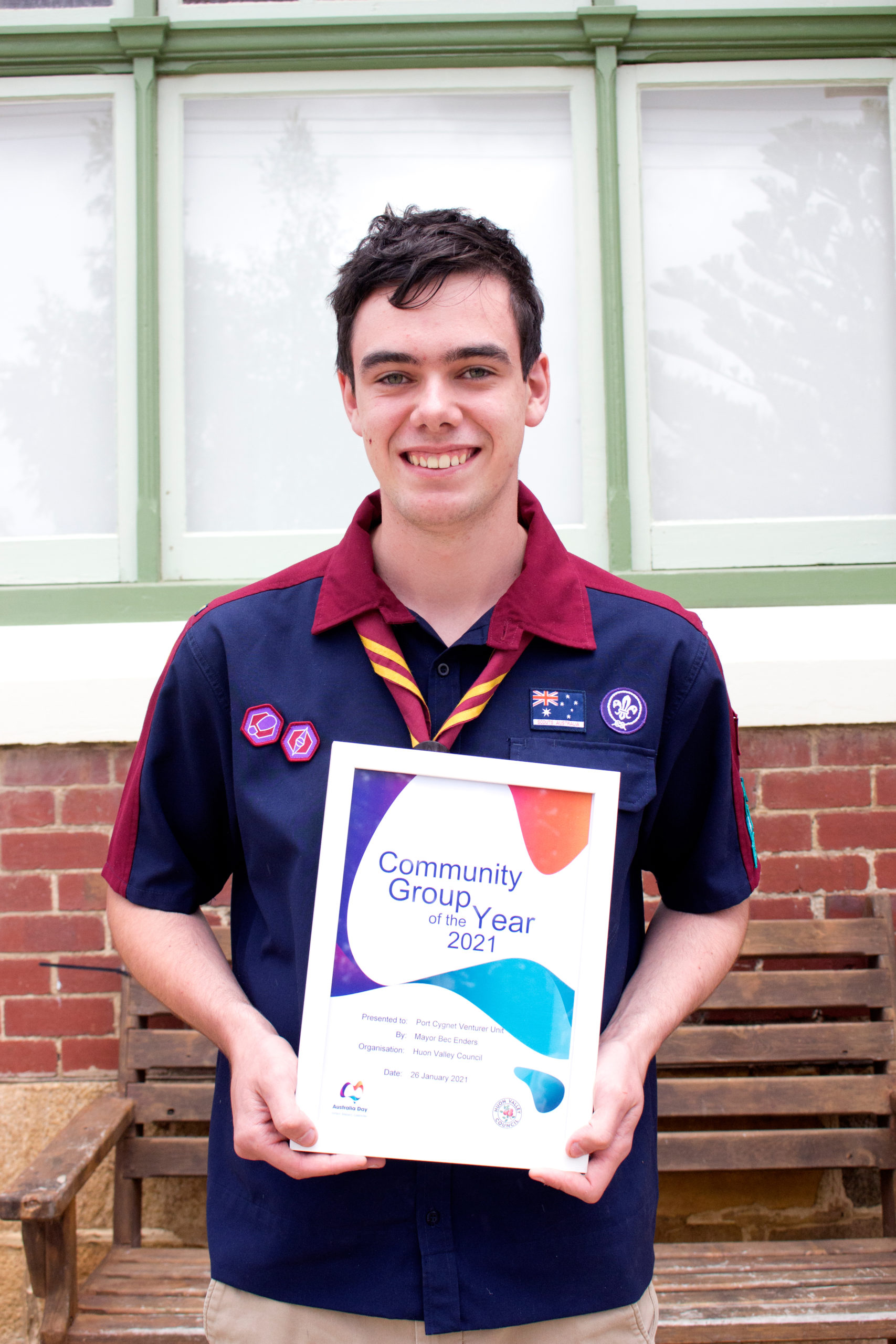 A person in a Scouts uniform holds a Community Group of the Year 2021 award. They are smiling and standing in front of a window and brick wall. The award has colorful graphics and a small Scouts logo.