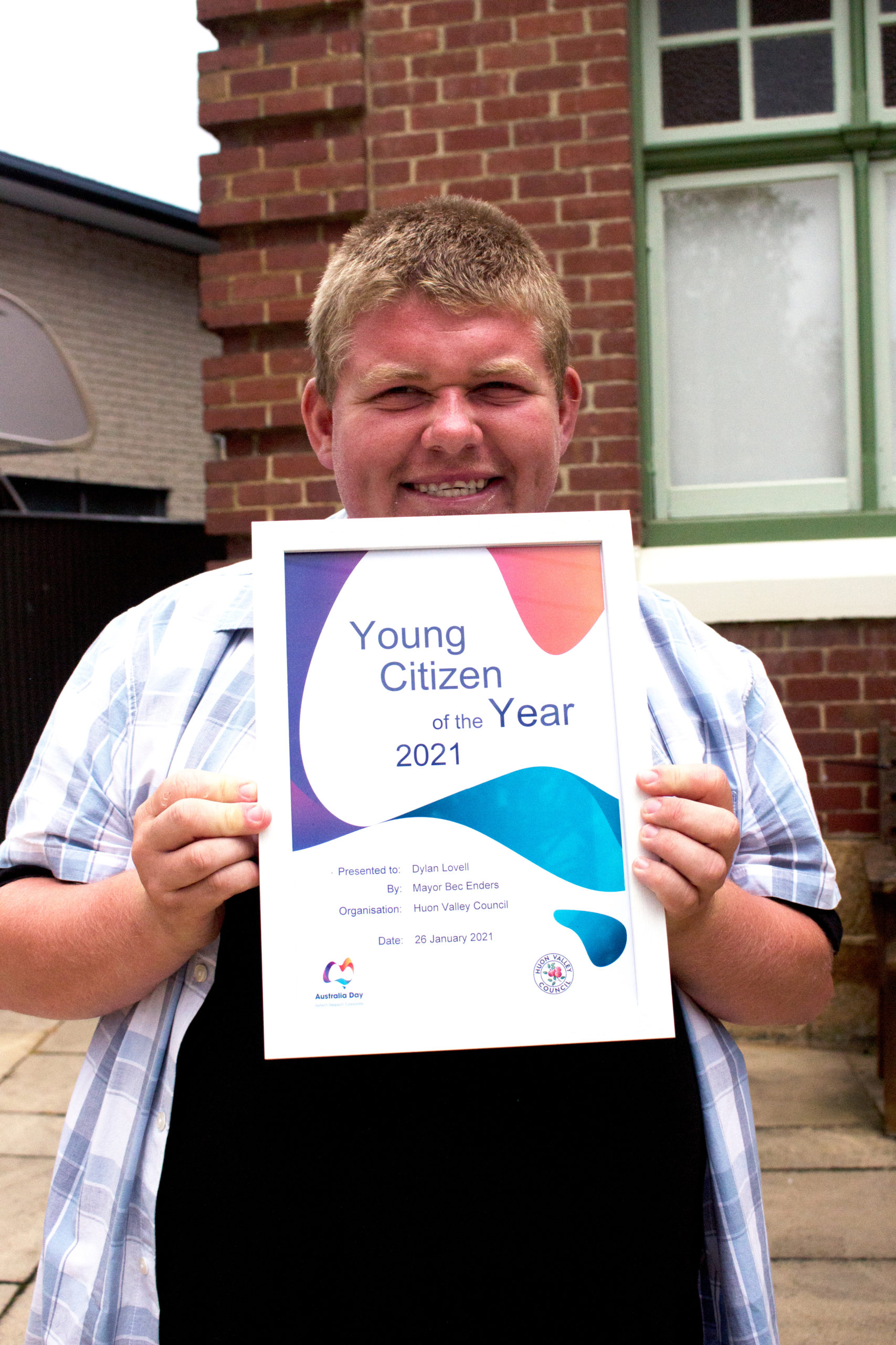 A person smiling while holding a framed certificate titled Young Citizen of the Year 2021. They are wearing a blue checkered shirt and standing in front of a brick building with a window.